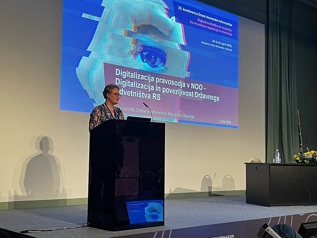 Saša Levstik na predavateljskem odru s projekcijo naslovne teme v ozadju.