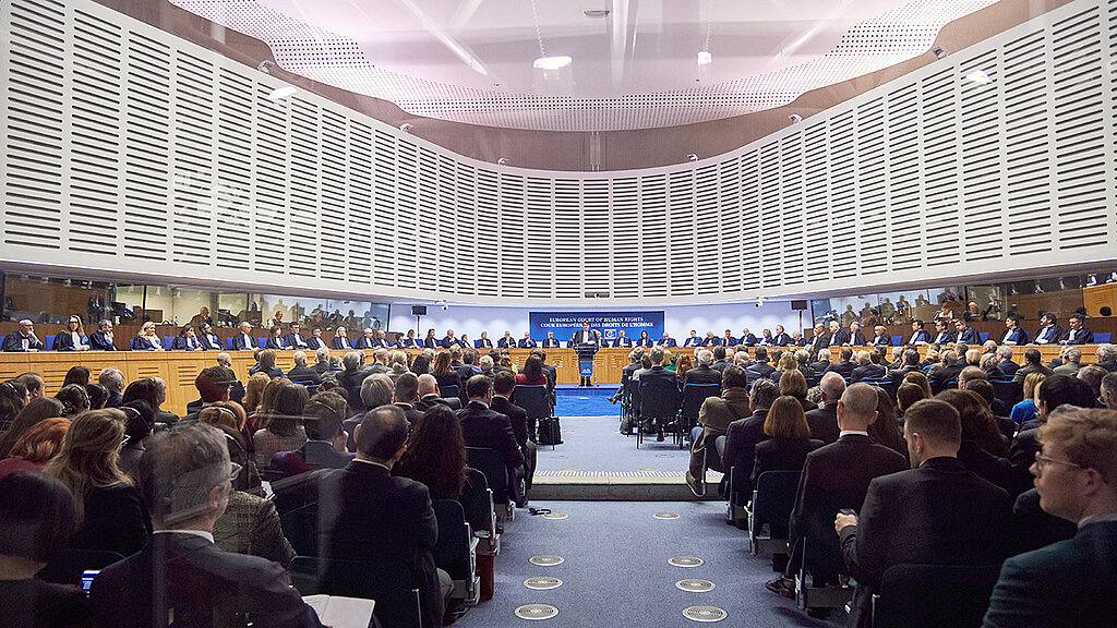 Polno zasedena glavna dvorana Evropskega sodišča za človekove pravice s prehodom v sredini, ki vodi do prostora za sodniški svet. 
