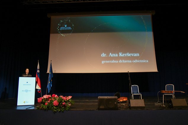 Nagovor generalne državne odvetnice Ane Kerševan, oder, govorniški pult desno, v sredini platno z imenom govorke, desno na odru dva stola, kitara